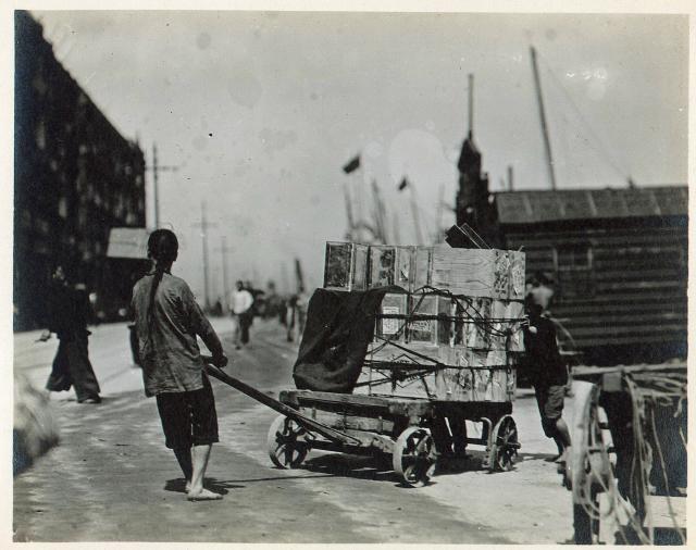 A person pulling a cart loaded with boxes. Docks and ships in the background