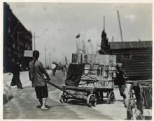 A person pulling a cart loaded with boxes. Docks and ships in the background