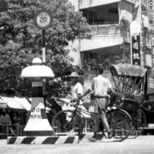A cyclist and a rickshaw puller, waiting to cross Nathan Road at the junction with Austin Road in the 1950s